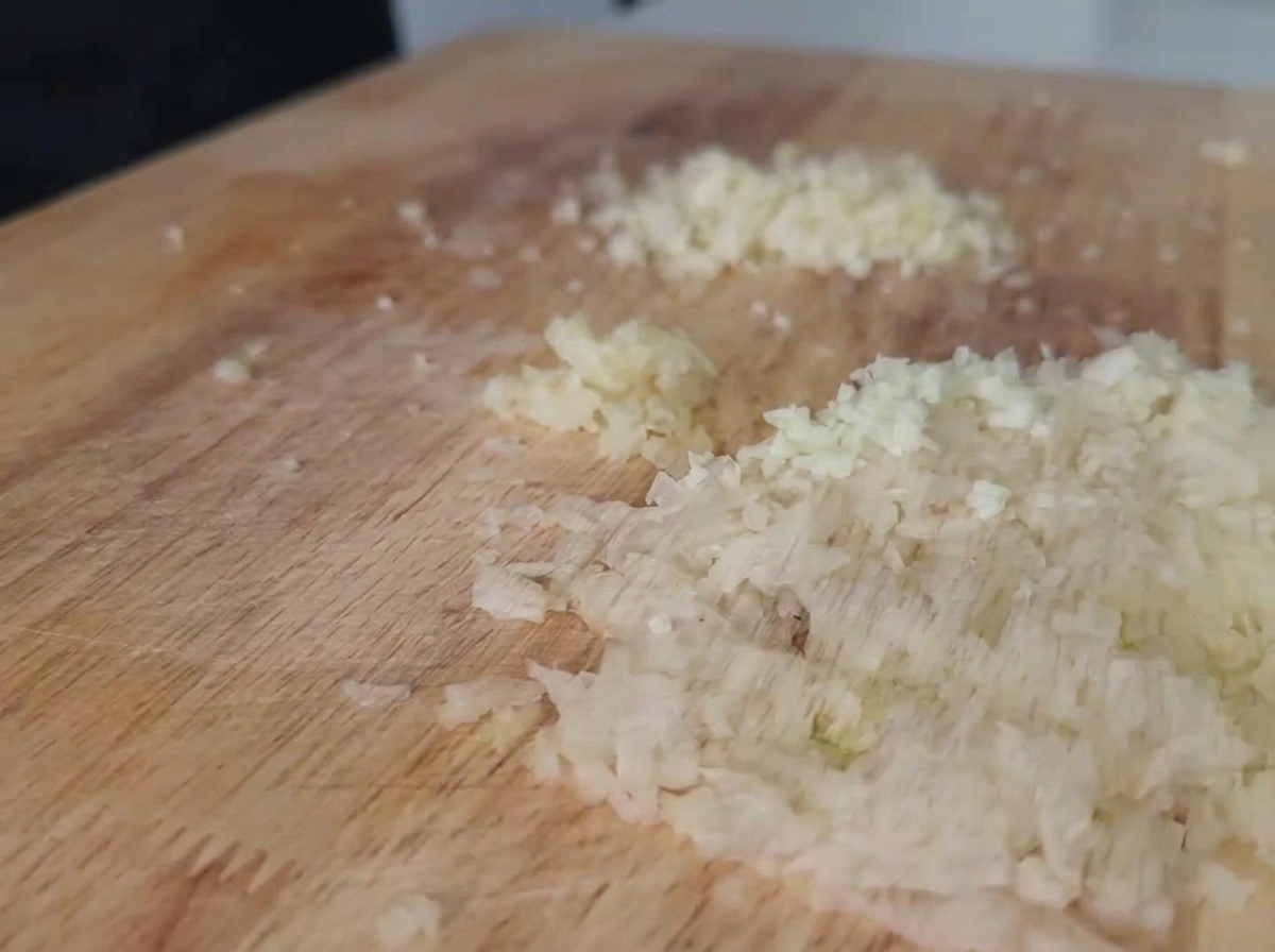 Finely minced white onions and garlic spread out on a light-colored wooden cutting board.