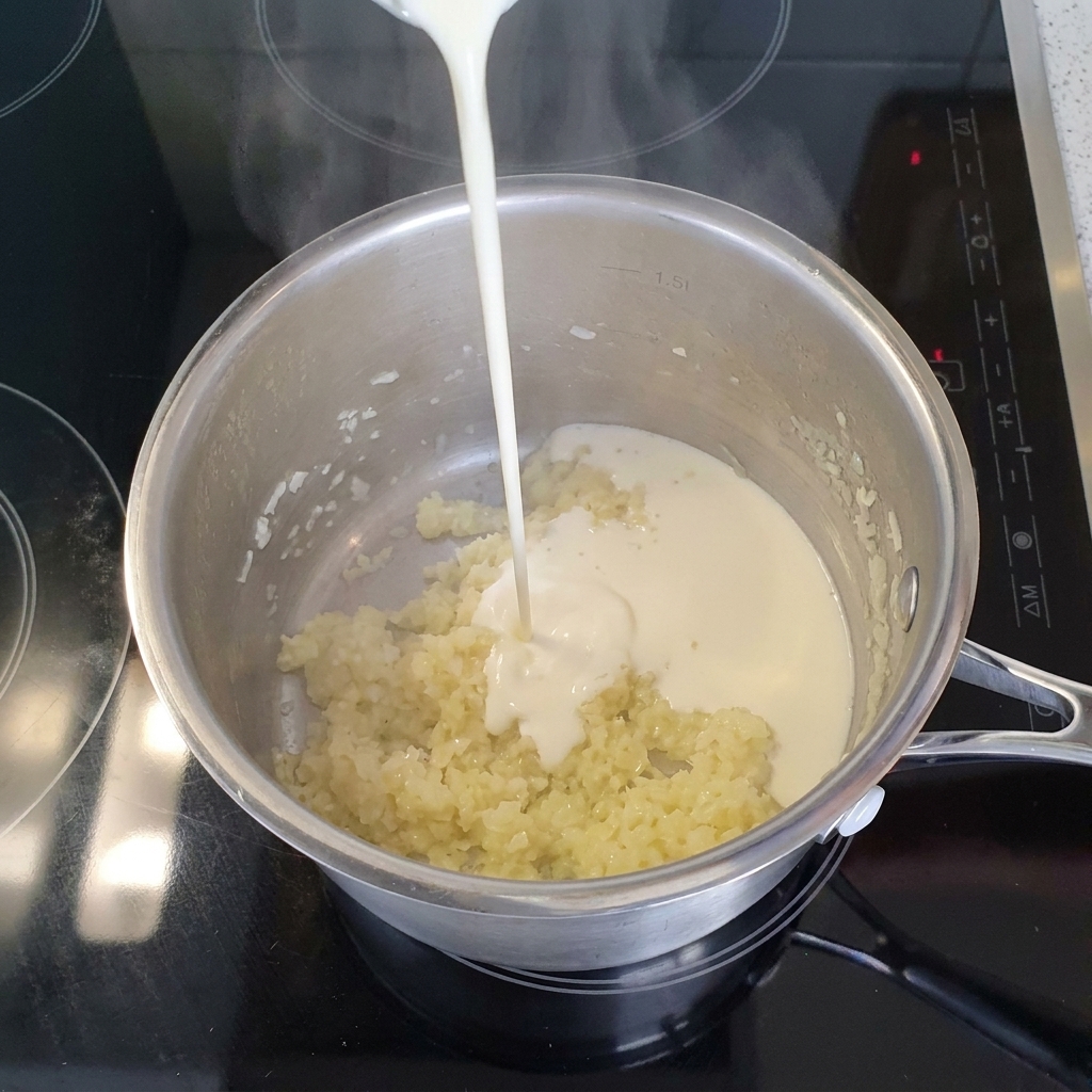 A stream of liquid light cream being poured into a stainless steel pot filled with cooked minced onions and garlic.