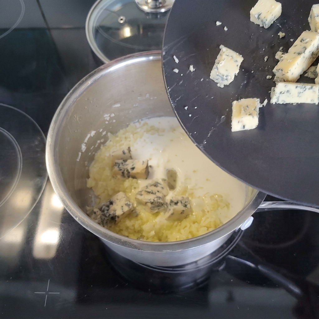 Chunks of blue cheese sliding off a tilted black plate into a saucepan containing cream and sautéed aromatics.
