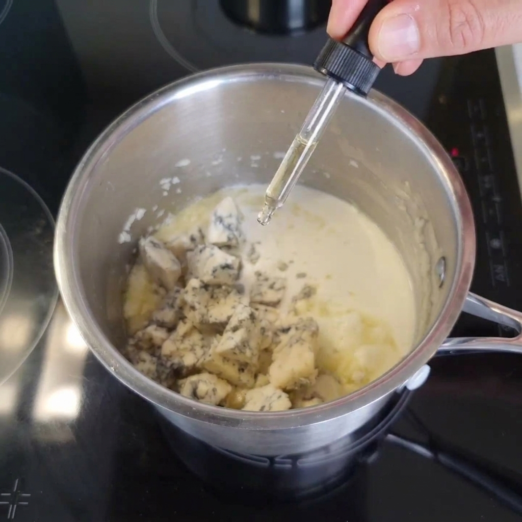 A hand using a glass dropper to add dark Worcestershire sauce into a metal pot filled with light cream and blue cheese chunks.