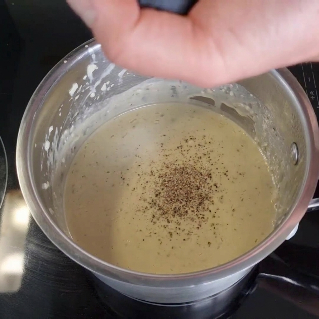 A hand grinding fresh black pepper from a mill into a creamy sauce inside a stainless steel pot on a black stovetop.
