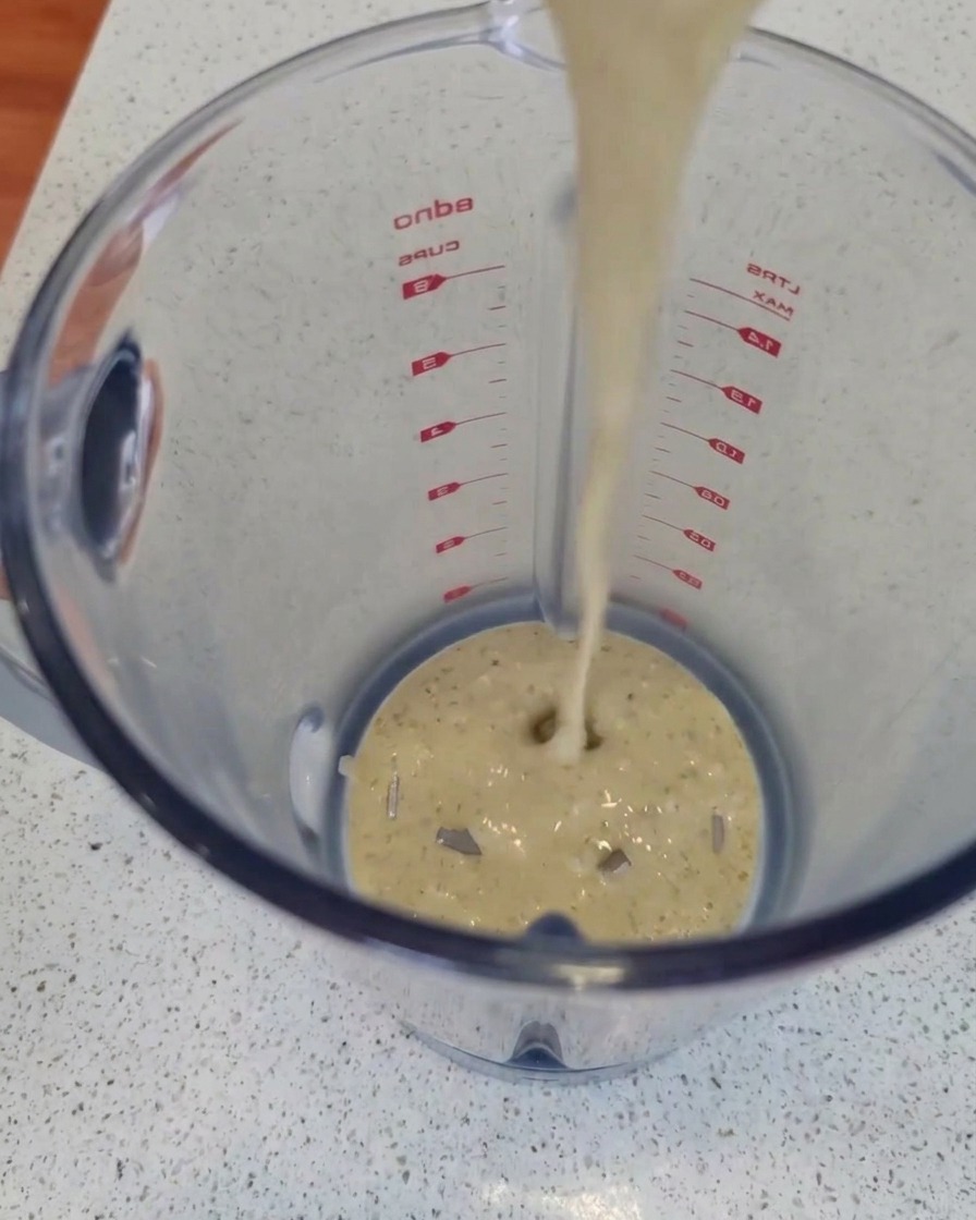 A stream of creamy, flecked blue cheese sauce being poured from above into a clear plastic blender jar on a kitchen countertop.