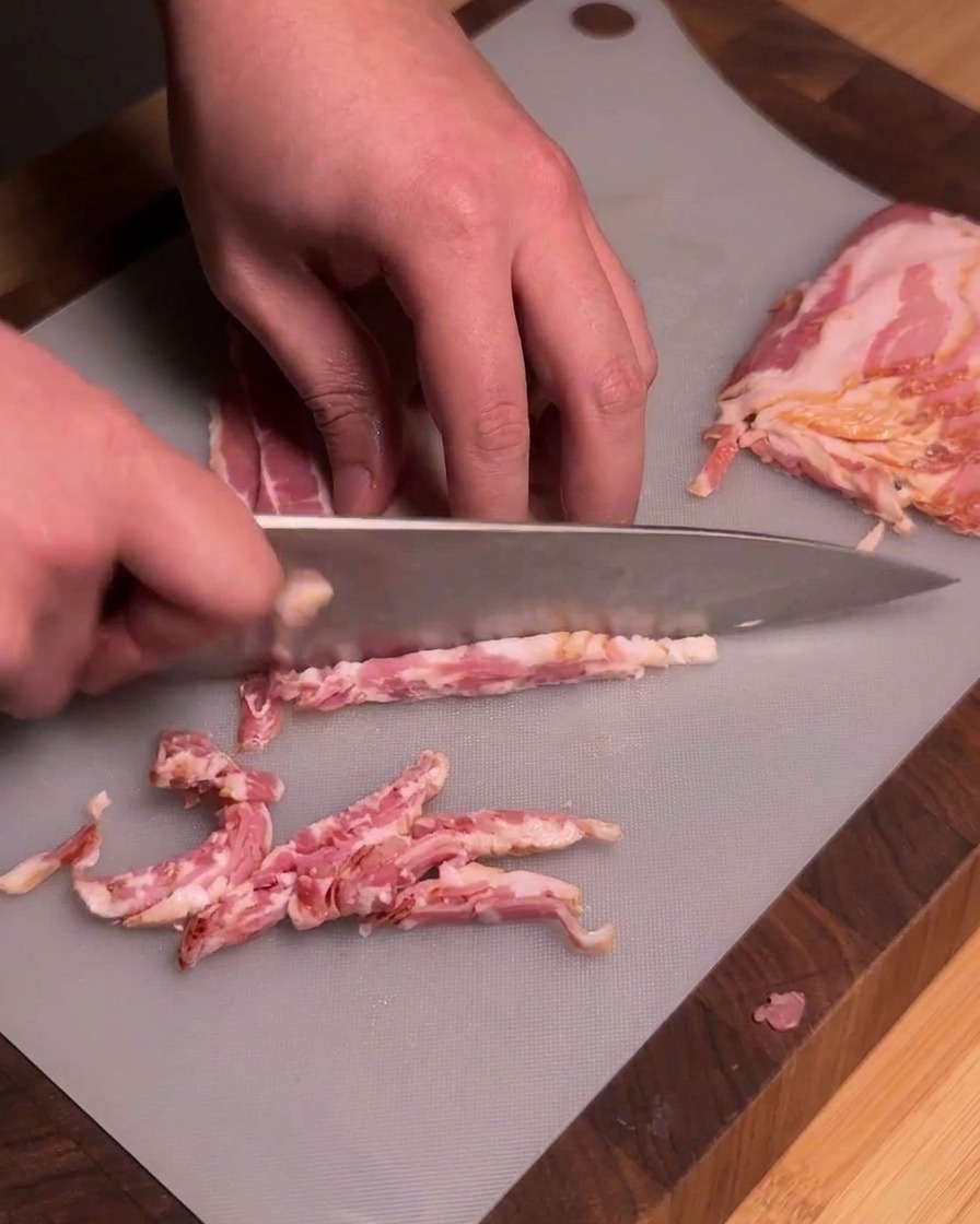 A chef using a sharp kitchen knife to slice raw strips of bacon into uniform pieces on a gray cutting board.