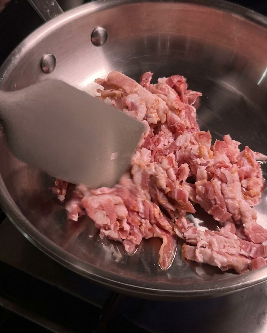 Diced raw bacon pieces being tossed and cooked with a metal spatula inside a heated stainless steel skillet.