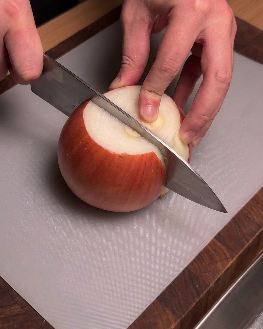 Hands holding a chefs knife to slice through a peeled yellow onion on a gray cutting board.