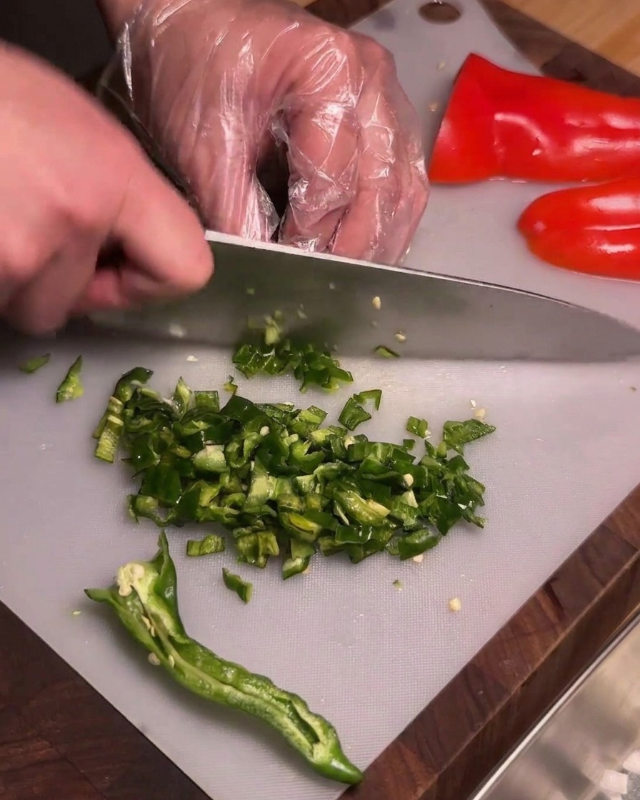A person wearing clear plastic gloves finely dicing deseeded green wrinkled chili peppers on a cutting board.