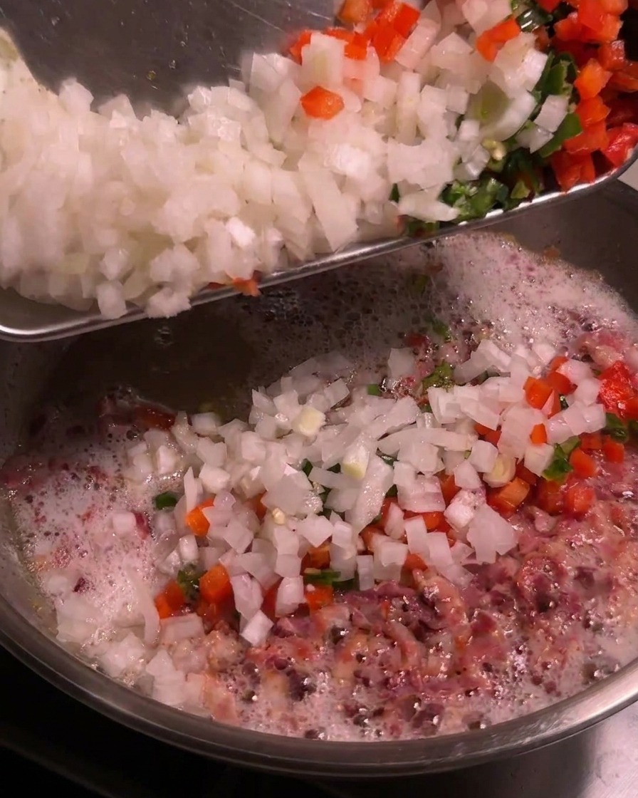 A tray of diced white onions and chopped colorful chili peppers being scraped into a bubbling skillet of rendered bacon fat.