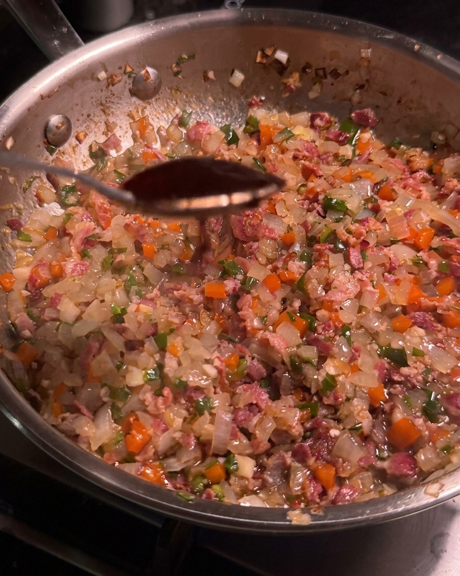A spoonful of dark chipotle pepper sauce held over a skillet filled with bacon and vegetable mixture.