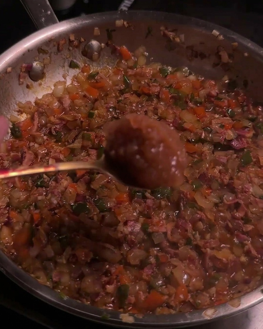 A spoon with applesauce being added to a caramelized bacon and pepper mixture in a pan.