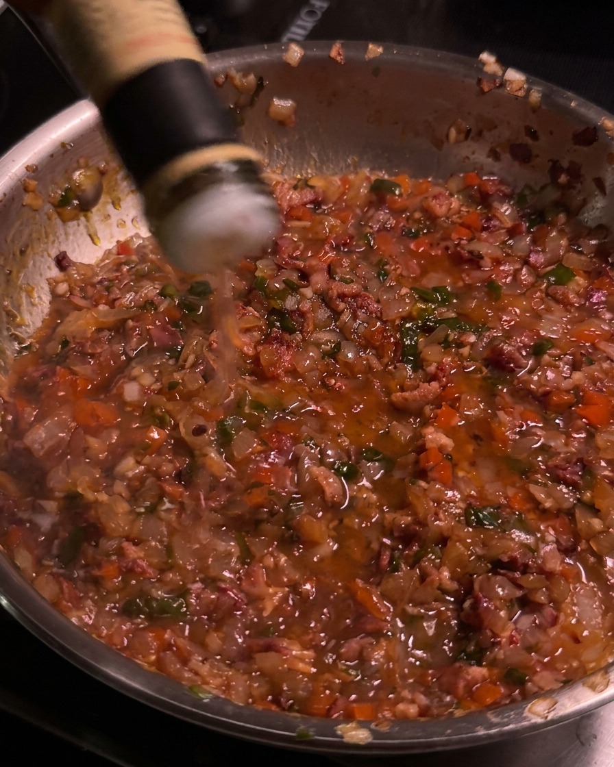 Apple cider vinegar being poured from a bottle into the simmering bacon jam skillet.