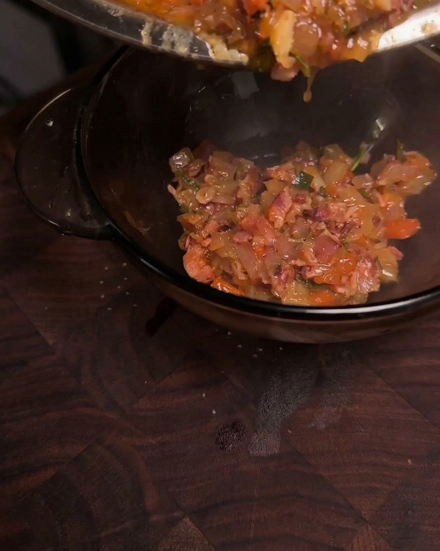 Thick and glossy caramelized bacon jam being poured from a pan into a clear glass bowl.