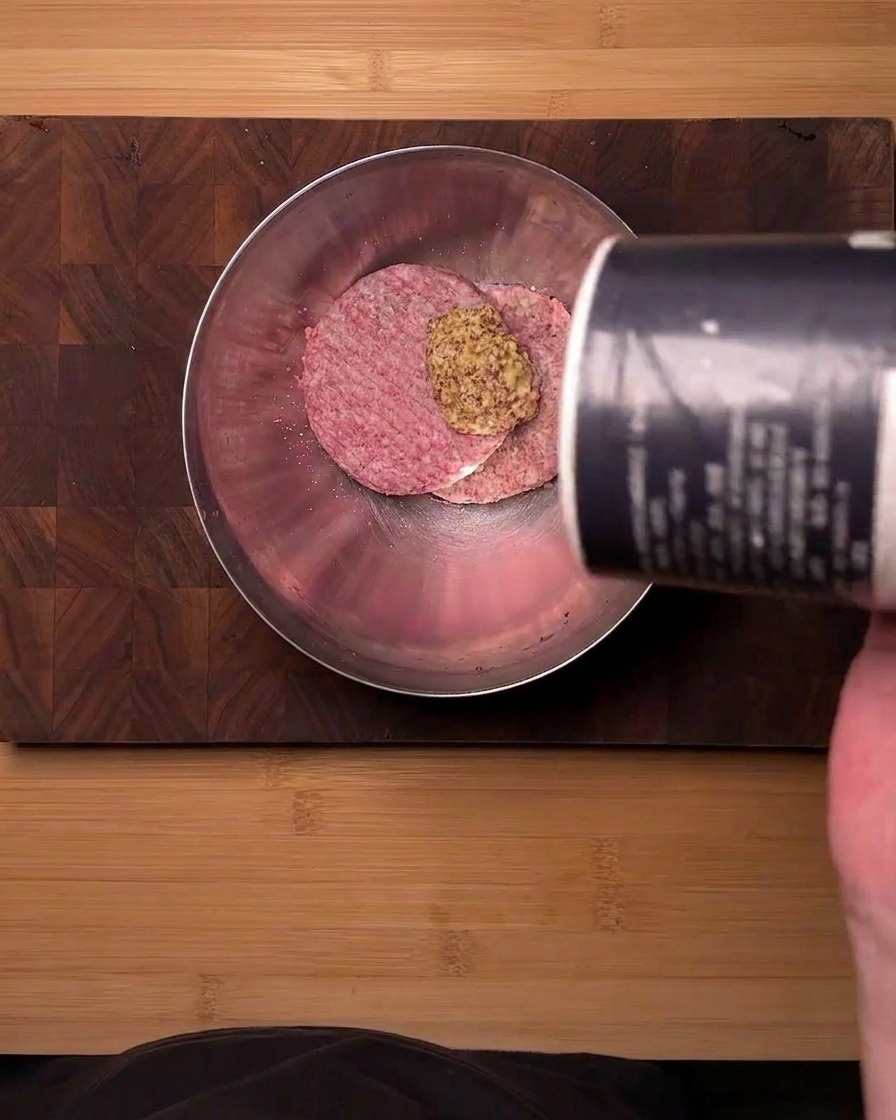 A top-down view of raw ground beef in a stainless steel bowl being seasoned with mustard powder and salt poured from a container.