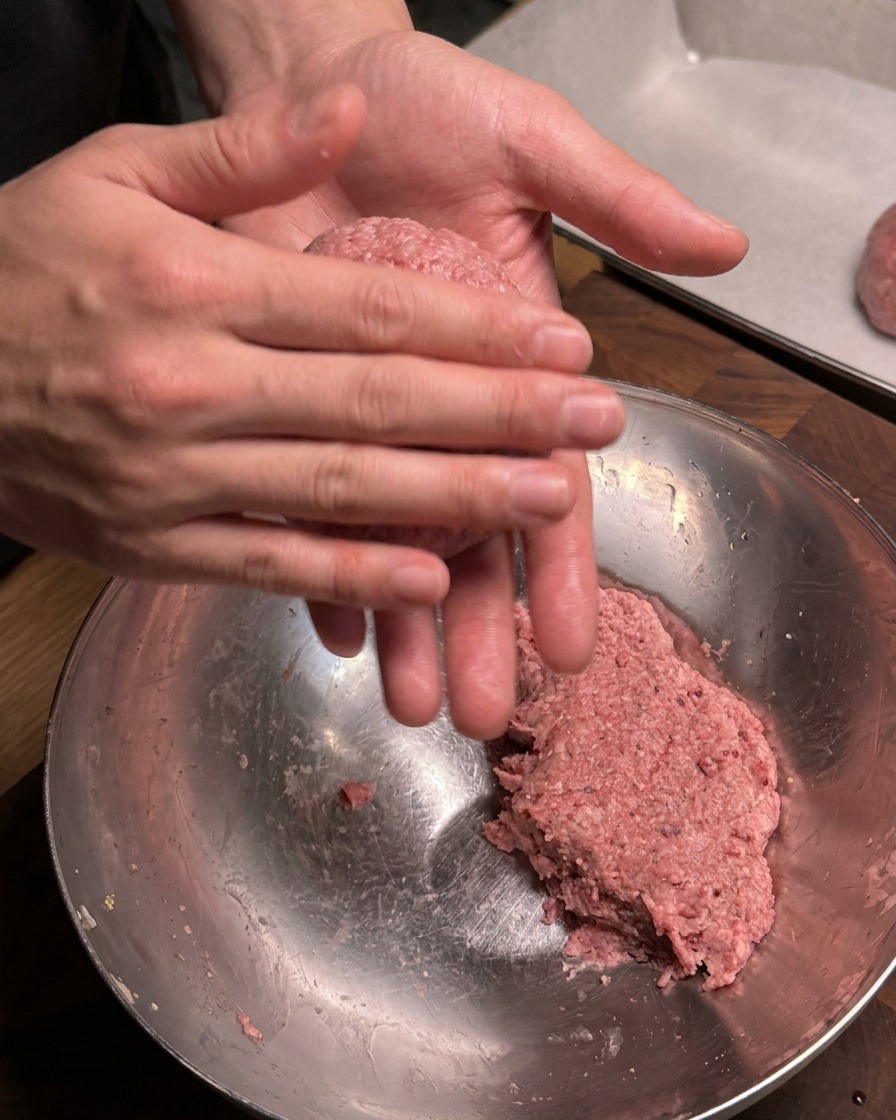A close-up view of hands firmly rolling and shaping a portion of seasoned raw ground beef into a round meatball over a metal mixing bowl.