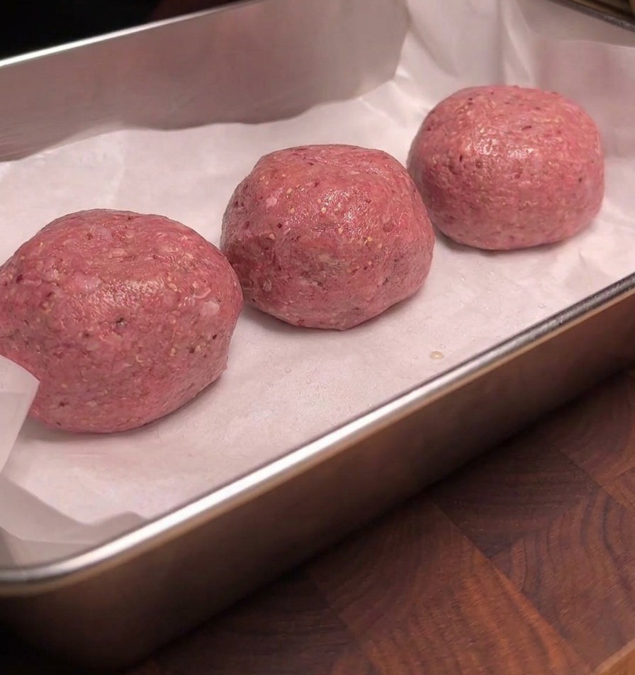 Three raw beef meatballs arranged neatly on white parchment paper inside a metal tray, their surfaces glistening uniformly with a coating of liquid smoke.