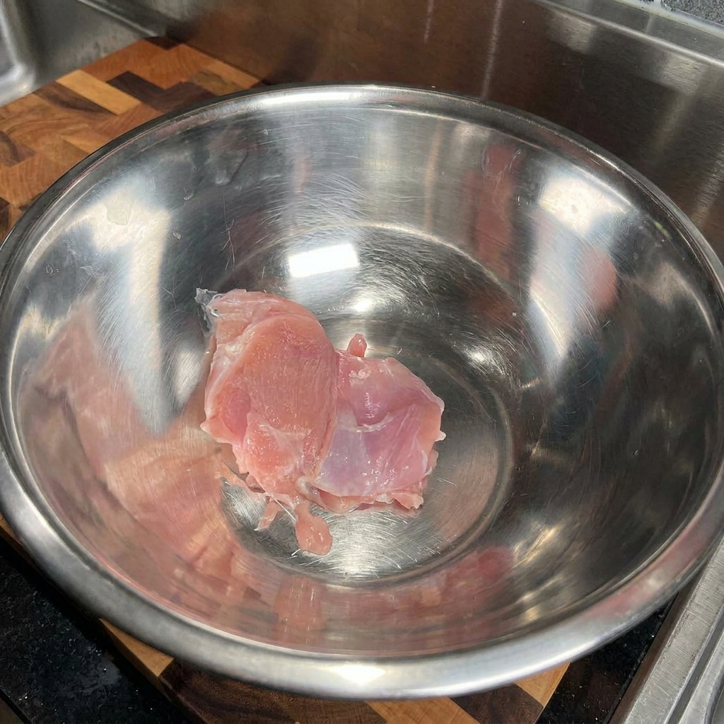 Raw chicken thigh placed inside a large, empty stainless steel mixing bowl.