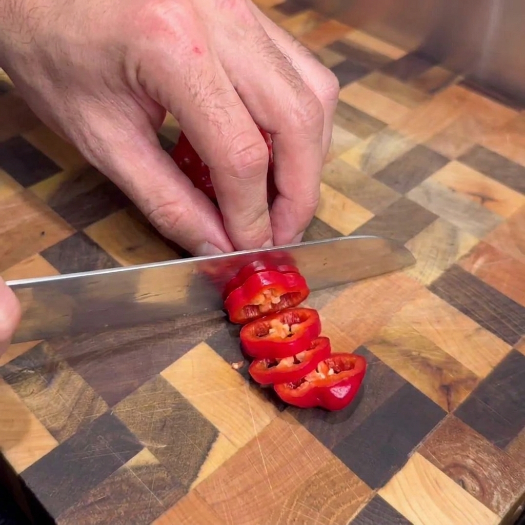 A person using a kitchen knife to slice fresh red hot peppers into rounds on an end-grain wooden cutting board.