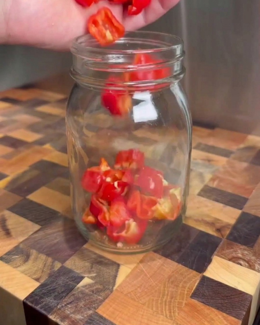 Chopped red peppers being dropped into a clear glass jar on a wooden cutting board.