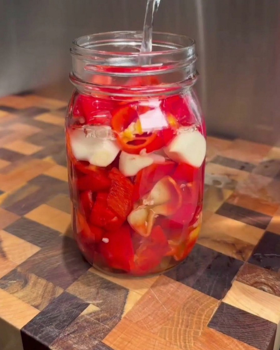 Clear salt brine being poured from a measuring cup into a glass mason jar filled with chopped red peppers and garlic cloves.