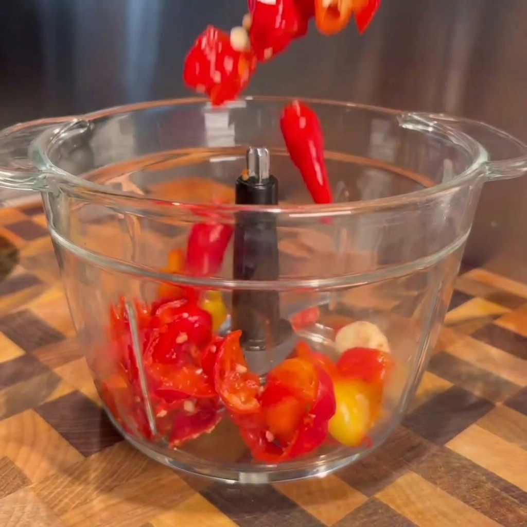 Drained red fermented peppers and garlic cloves falling into a clear food processor bowl.
