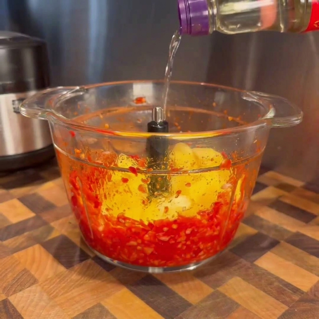 Clear liquid being poured from a bottle into a glass food processor bowl containing blended red chilies and bright yellow mango chunks.