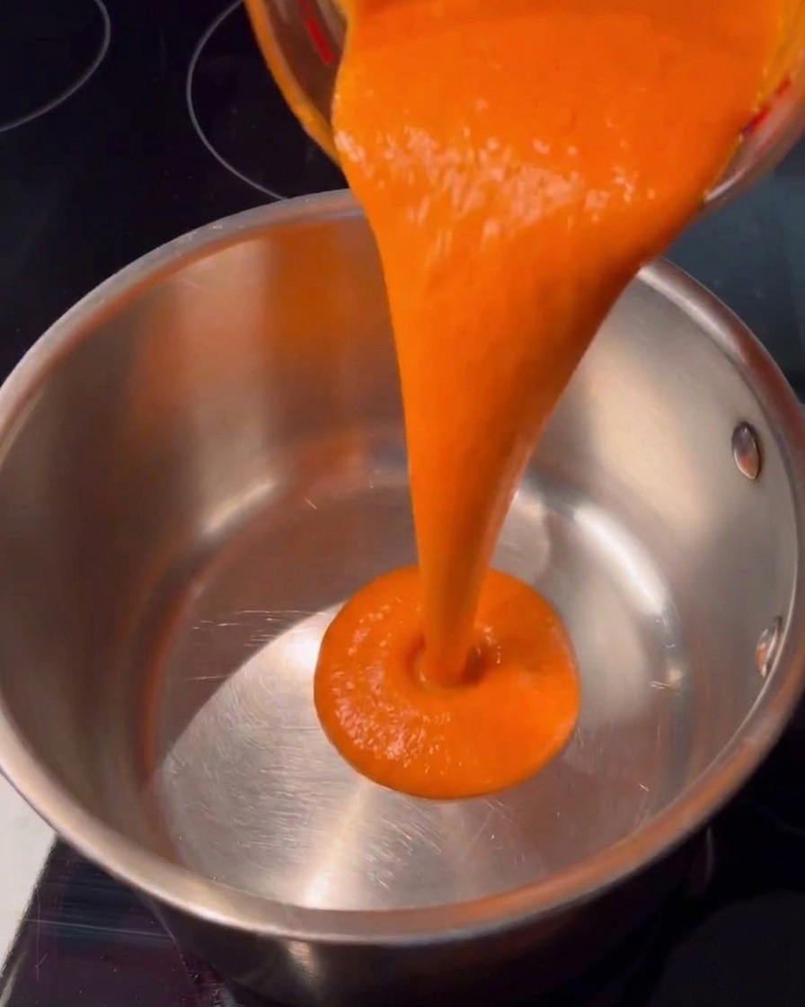 Bright orange hot sauce pouring seamlessly from a glass cup into a clean stainless steel saucepan on a dark stovetop.