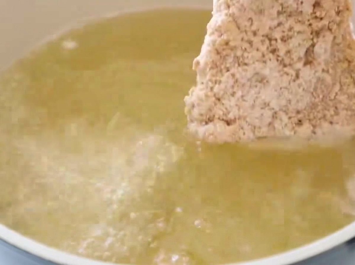 A breaded chicken fillet being submerged into a pot of shimmering, hot bubbling cooking oil for deep frying.