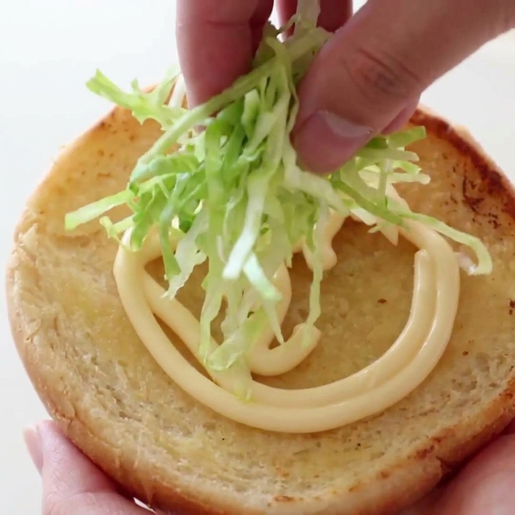 A hand placing a pile of crisp, finely shredded green lettuce onto a toasted burger bun layered with a swirl of creamy mayonnaise.