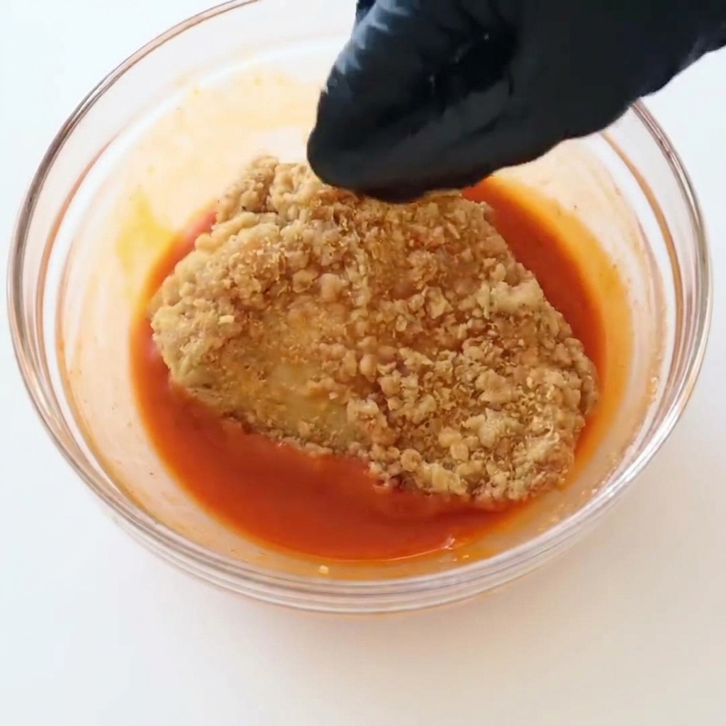 A hand wearing a black glove lowering a crispy, golden fried chicken fillet into a clear bowl filled with bright red buffalo sauce.