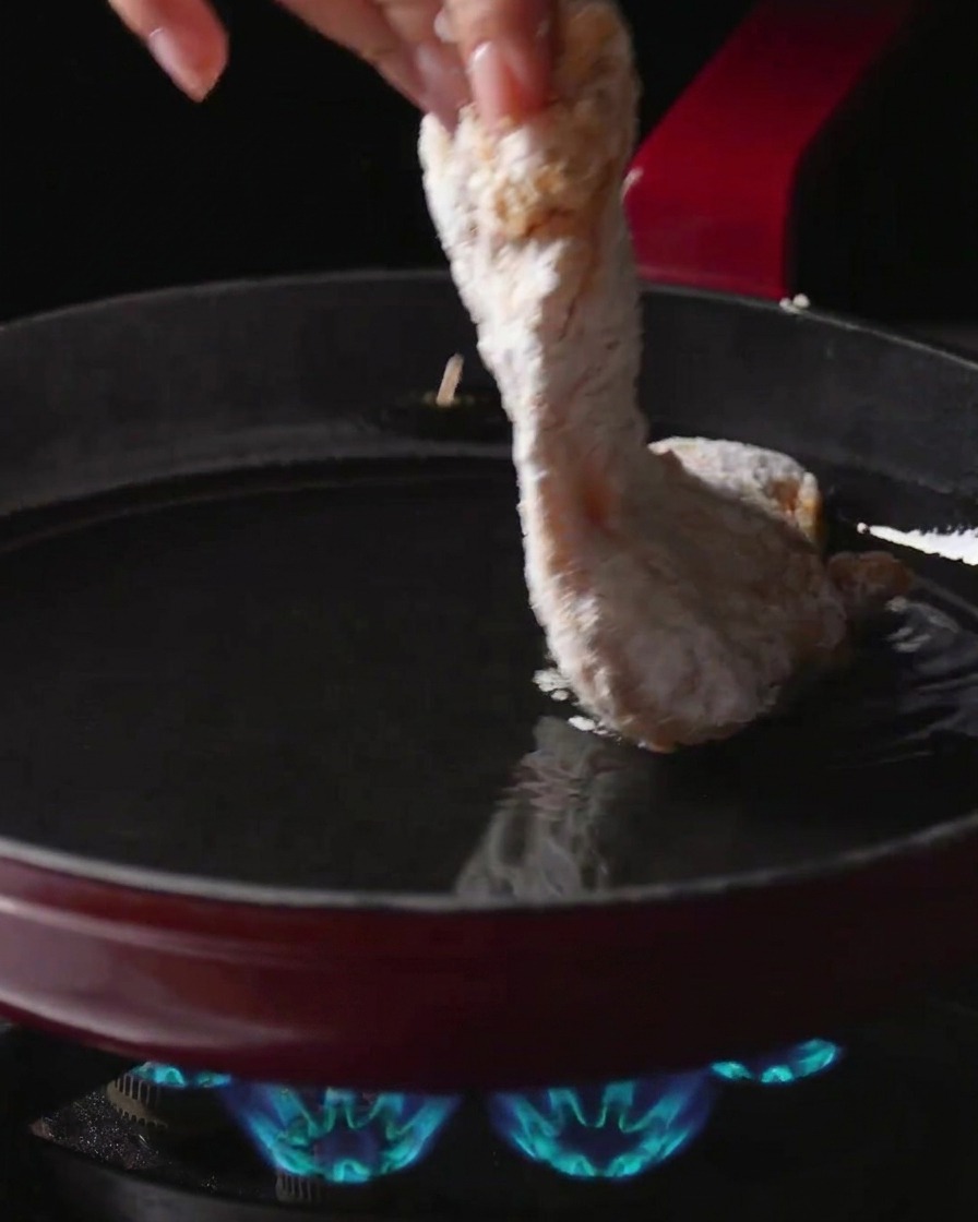 A hand placing a flour-coated chicken thigh into a pan of hot oil over a lit gas burner.