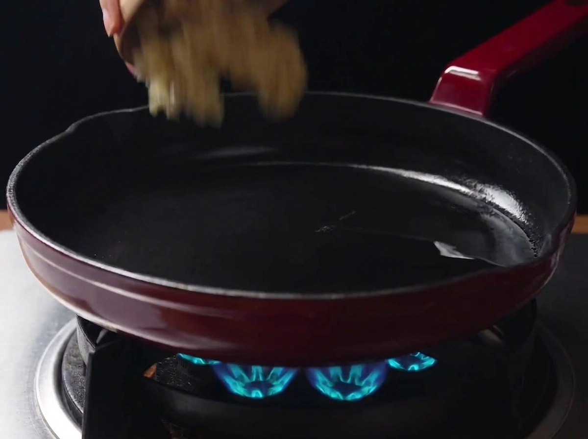 Minced garlic being added from a small bowl into a hot frying pan with oil over a lit gas stove.