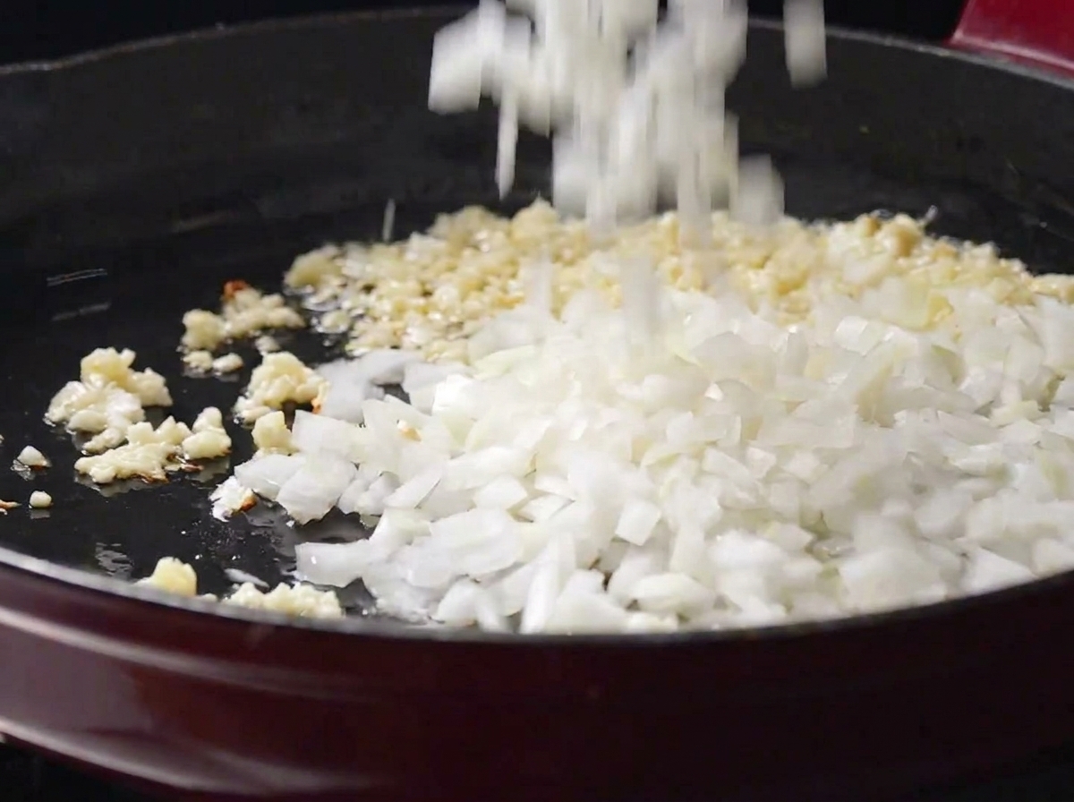 Diced white onions being added to a frying pan with sizzling minced garlic.