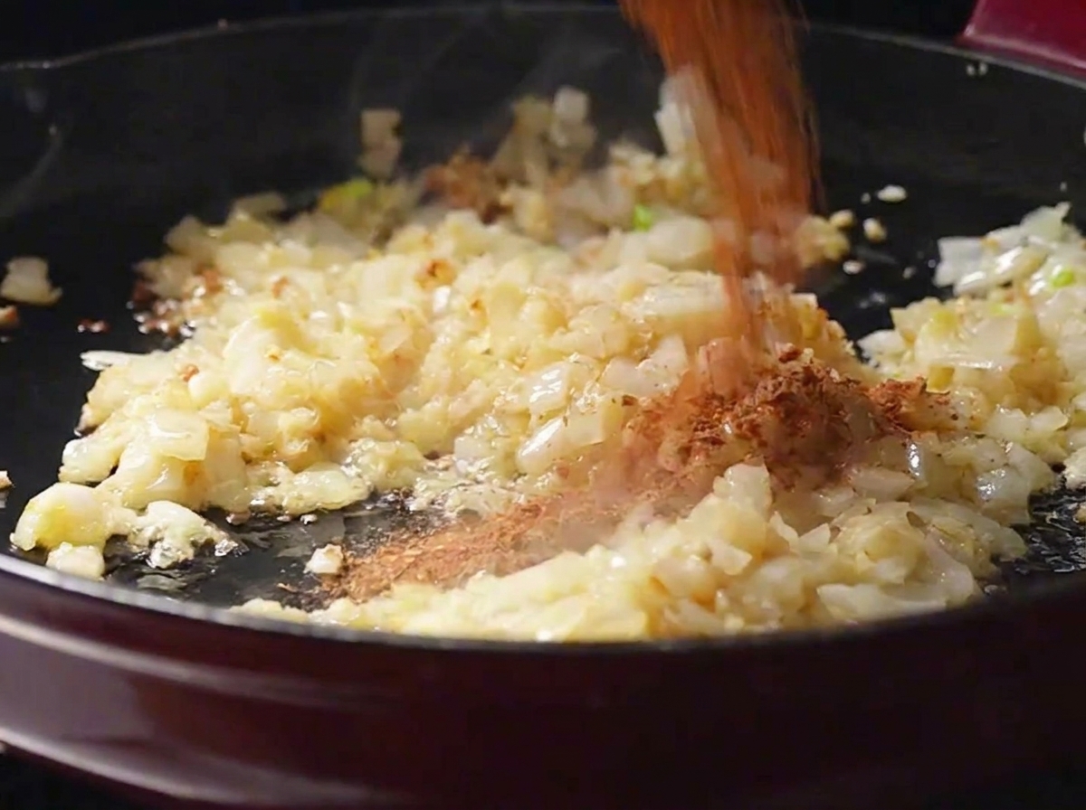 Homemade red-brown Cajun spice powder being poured over sautéed onions and garlic in a frying pan.
