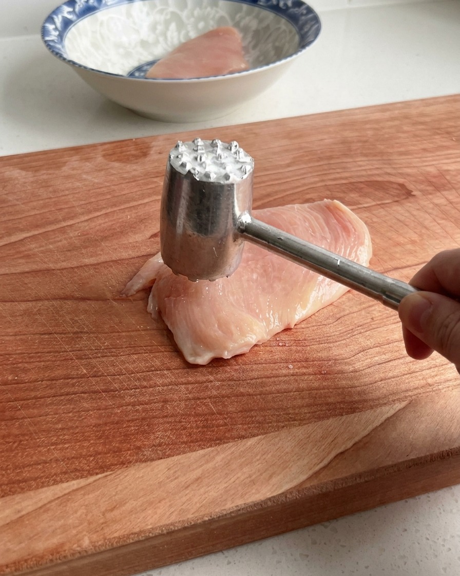 A hand using a metal meat mallet to gently pound a raw chicken breast on a large wooden cutting board.