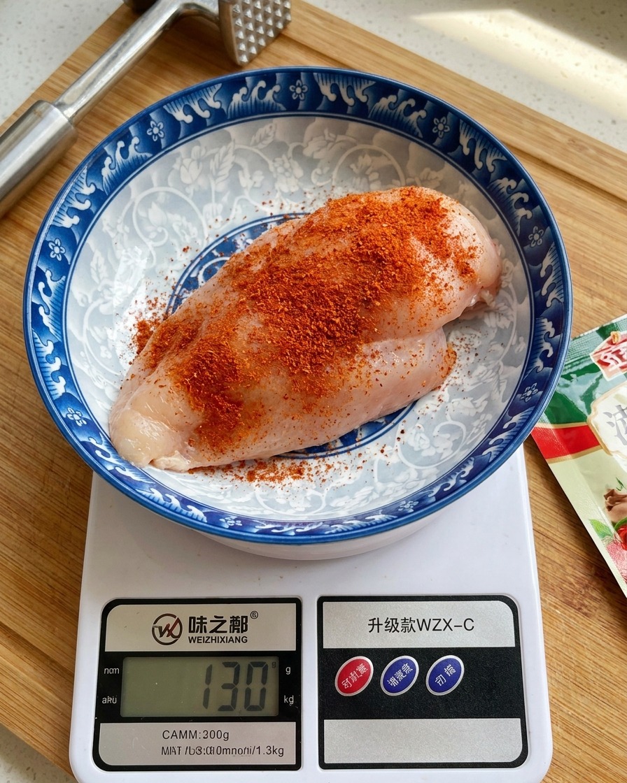 A piece of raw chicken breast in a decorated ceramic bowl on a digital kitchen scale, heavily dusted with orange-red seasoning powder.