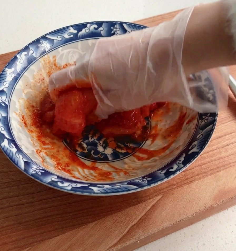 A gloved hand thoroughly coating and mixing a raw chicken breast in a thick red marinade mixture inside a patterned bowl.
