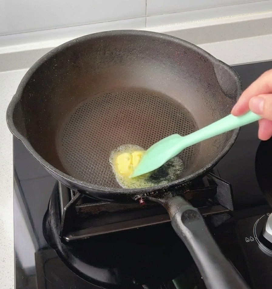A green silicone spatula melting a small pat of yellow butter in the center of a hot black pan.