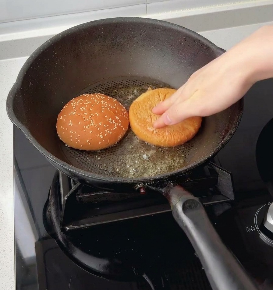 A hand pressing the top and bottom halves of a sesame burger bun face-down into melted butter inside a skillet.