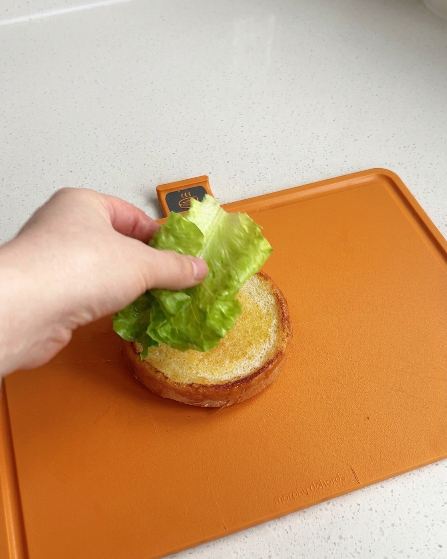 A hand placing a piece of clean, vibrant green lettuce leaf on top of a golden, butter-toasted burger bun base on an orange cutting board.