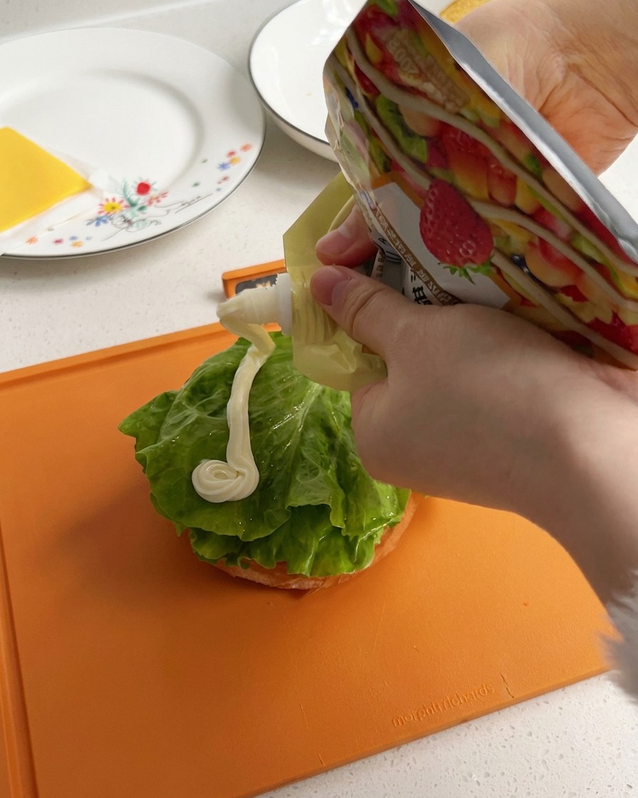 Creamy white mayonnaise being squeezed in a thick line across a green lettuce leaf on a burger bun base.