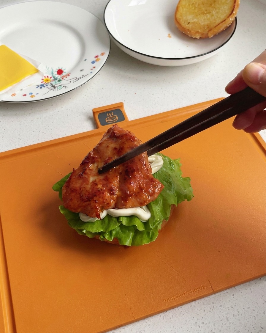 A hand using dark wooden chopsticks to transfer a glazed, deeply caramelized pan-fried chicken piece onto a burger stack.