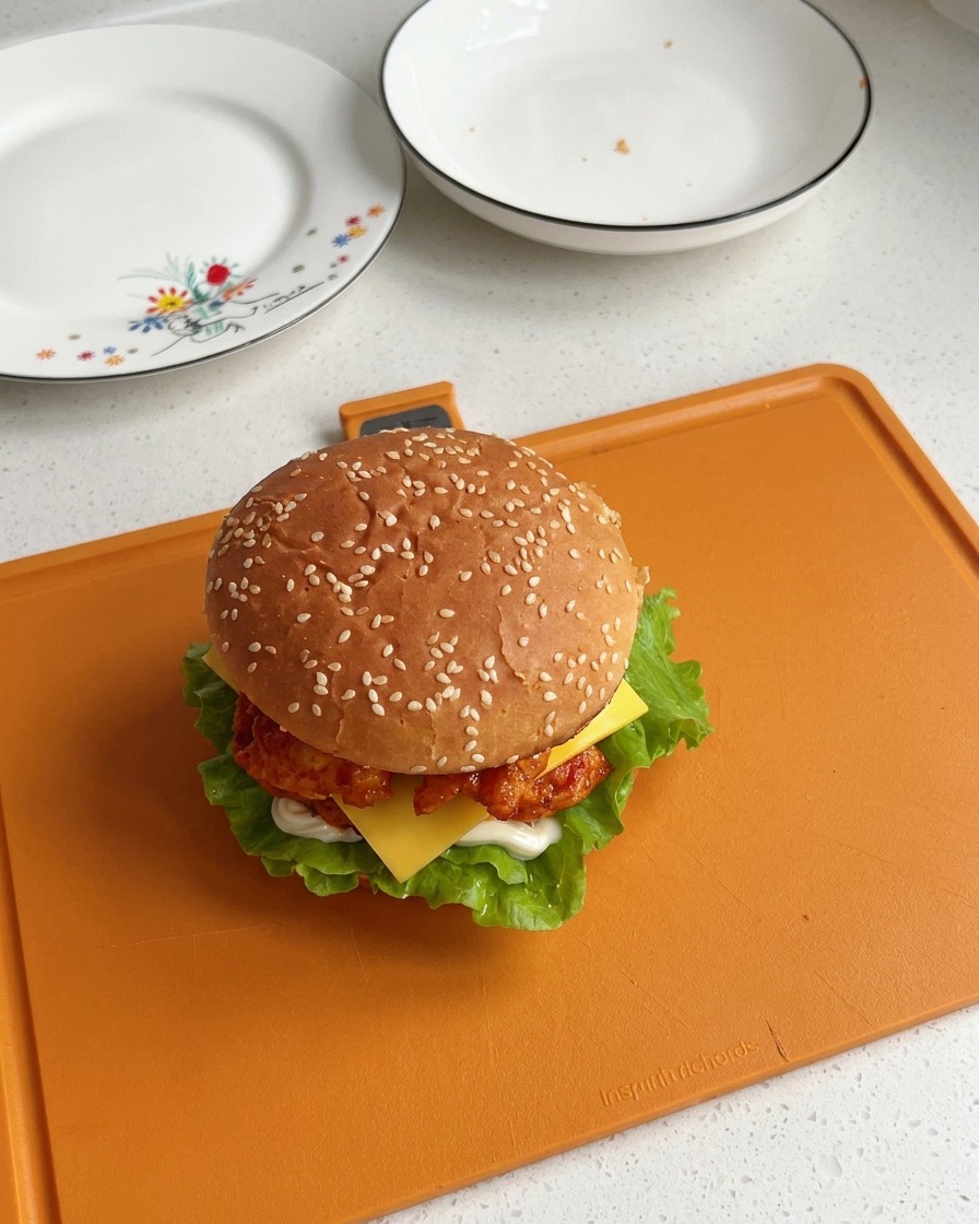 A fully assembled Orleans chicken burger topped with a sesame seed bun, showcasing visible layers of lettuce, chicken, and a melting cheese slice.