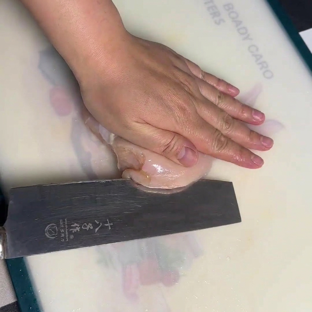 A hand holding a raw chicken breast flat on a white cutting board while carefully slicing it horizontally with a large chefs knife.