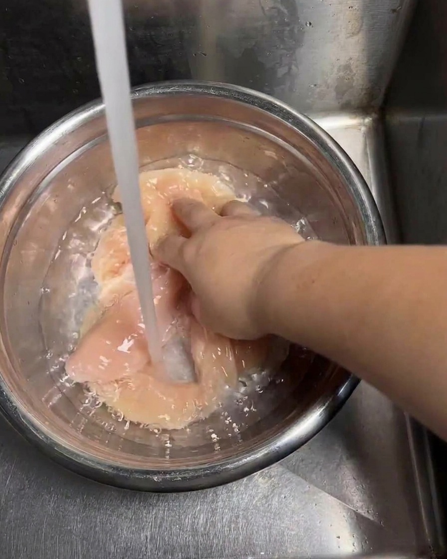 A stream of clean tap water filling a stainless steel bowl containing raw chicken cutlets as a hand moves the pieces around to rinse them.