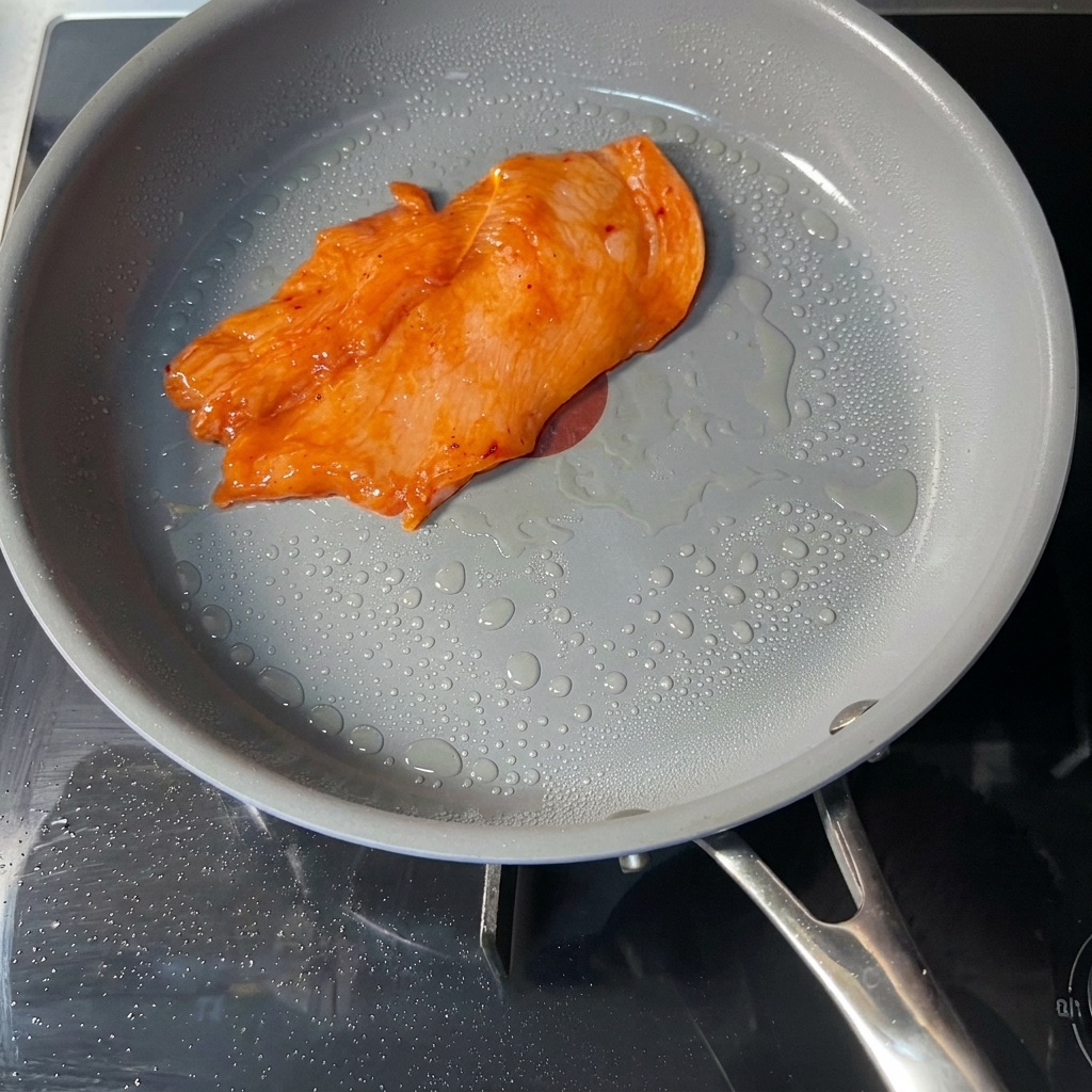 A single marinated orange-colored chicken cutlet cooking in a gray non-stick pan with visible droplets of oil on the surface.