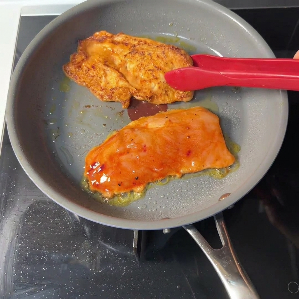 Red tongs flipping an orange marinated chicken cutlet in a gray skillet, revealing a golden-brown seared crust on the cooked side.