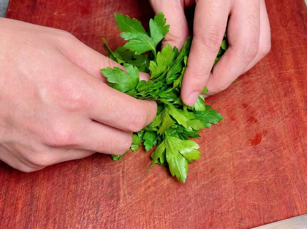 Two hands rolling up a bunch of fresh green flat-leaf parsley tightly on a red-brown wooden cutting board.