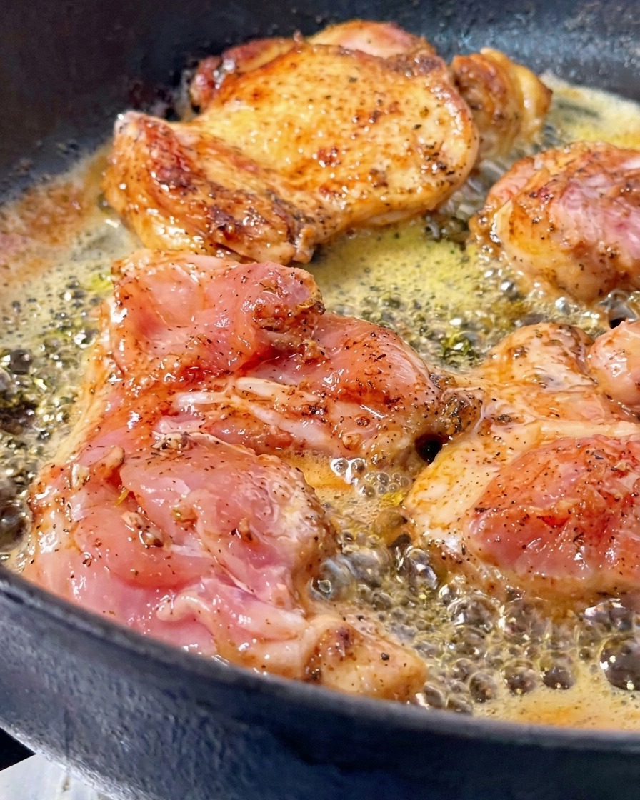 Chicken thighs being pan-seared in bubbling butter inside a cast-iron skillet, with one piece flipped over to reveal a crispy golden-brown crust.