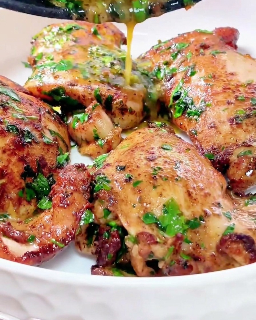 A stream of glossy golden butter sauce mixed with green herbs being drizzled from a pan over juicy, golden-brown chicken thighs in a clean white bowl.