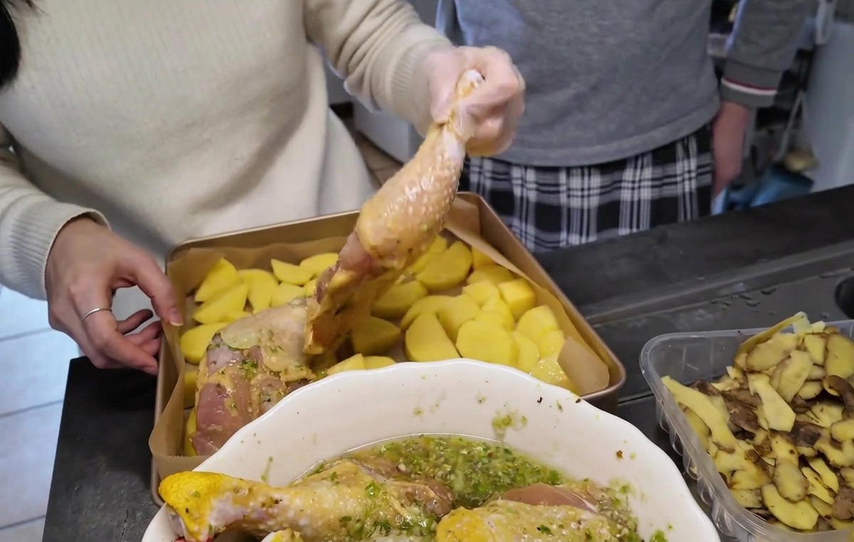 Raw marinated chicken legs being arranged skin-side up over a layer of chopped potatoes in a gold baking pan.