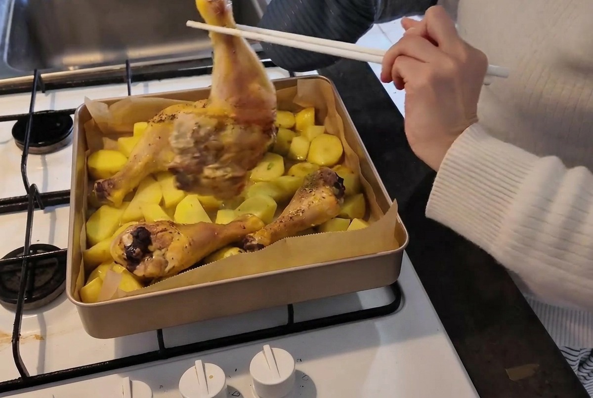 A person wearing an oven mitt uses chopsticks to flip a partially cooked chicken leg over in a hot baking pan filled with potatoes.